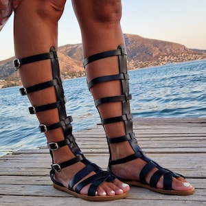 Op de afbeelding: Zwarte gladiatorsandalen van leer met meerdere banden en gespen. De sandalen hebben een platte zool en worden gedragen op een houten oppervlak.