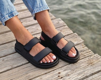 May include: Black sandals with two adjustable straps. The sandals have a thick sole and are worn with blue jeans. The image is taken on a wooden dock near the water.