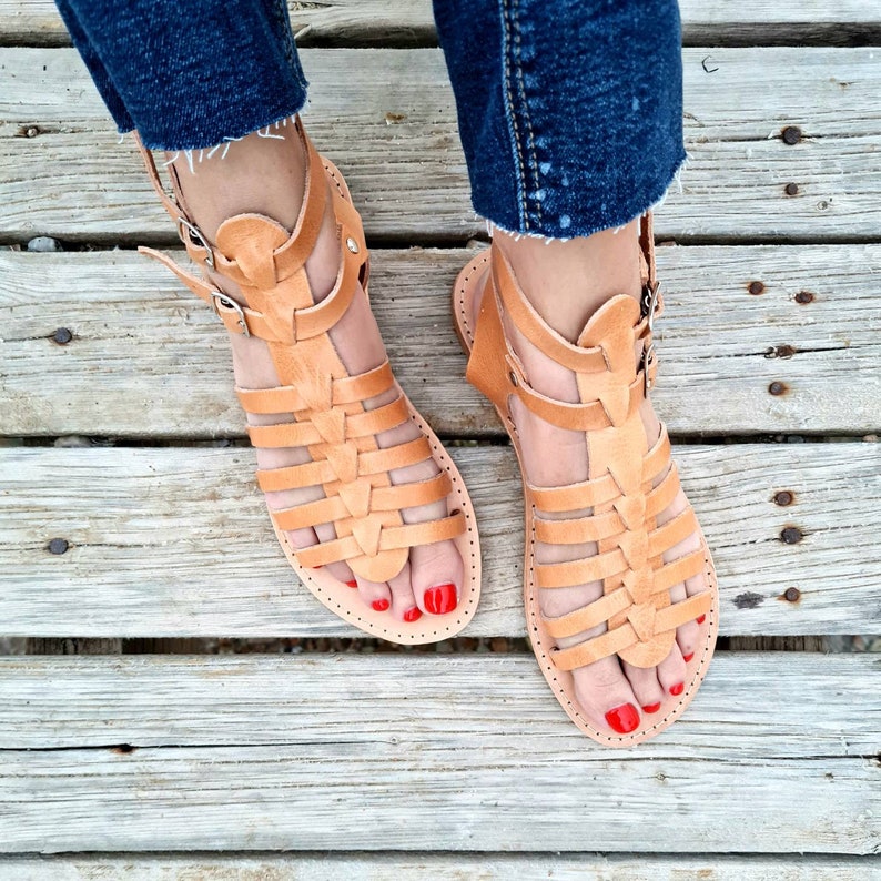 May include: A pair of tan leather gladiator sandals with a woven design. The sandals have a buckle closure at the ankle and red nail polish on the toes.