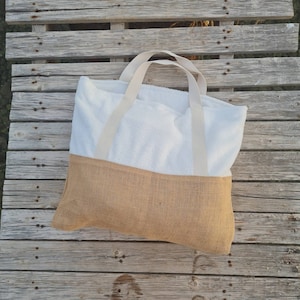 May include: A white beach tote bag with a brown burlap bottom and cream-colored straps. The bag is lying on a wooden surface.