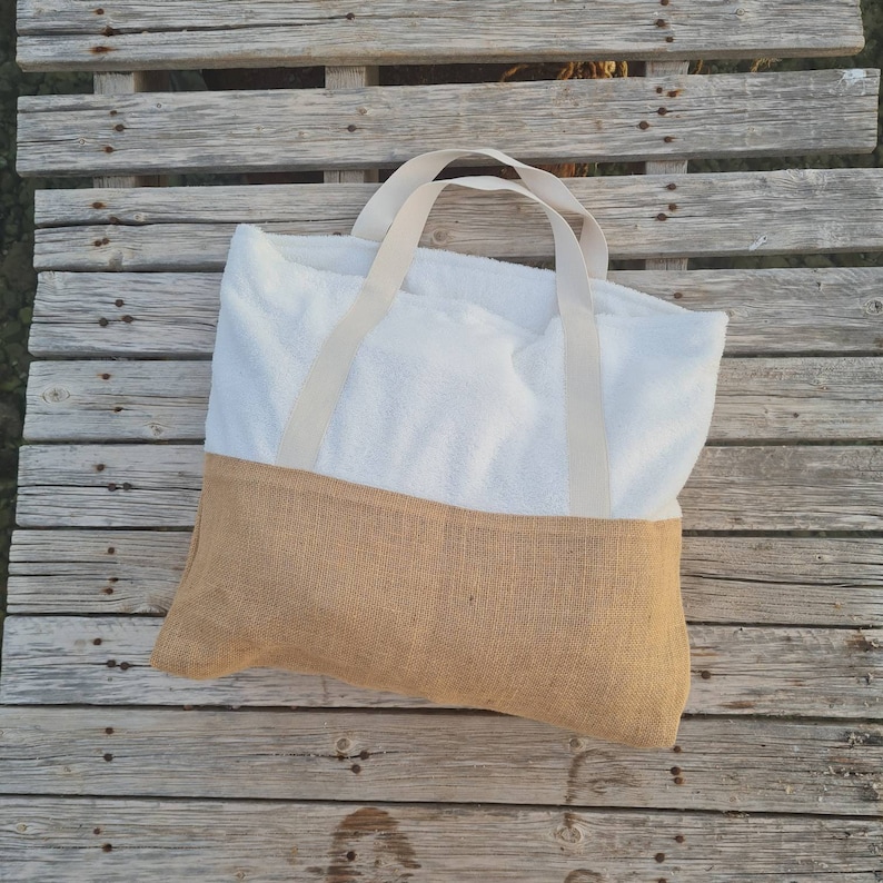 May include: A white beach tote bag with a brown burlap bottom and cream-colored straps. The bag is lying on a wooden surface.