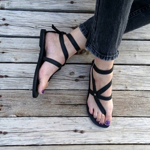 K&ouml;nnte beinhalten: Schwarze Ledersandalen mit mehreren Riemen und einer Zehenschlaufe. Die Sandalen werden auf einer Holzoberfl&auml;che getragen.