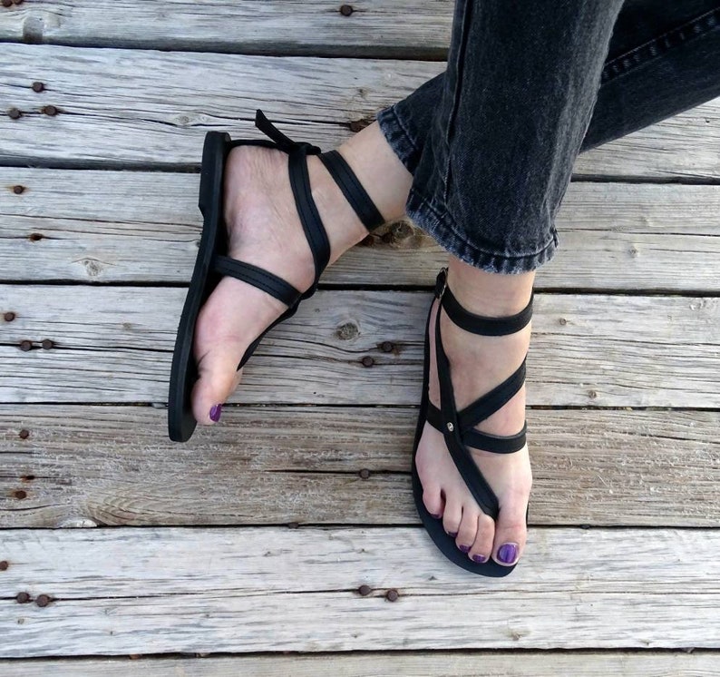 K&ouml;nnte beinhalten: Schwarze Ledersandalen mit mehreren Riemen und einer Zehenschlaufe. Die Sandalen werden auf einer Holzoberfl&auml;che getragen.
