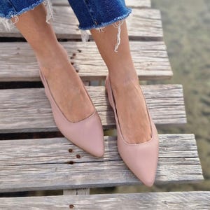 May include: A pair of blush pink leather slip-on shoes with pointed toes. The shoes are worn by a person standing on a wooden walkway.