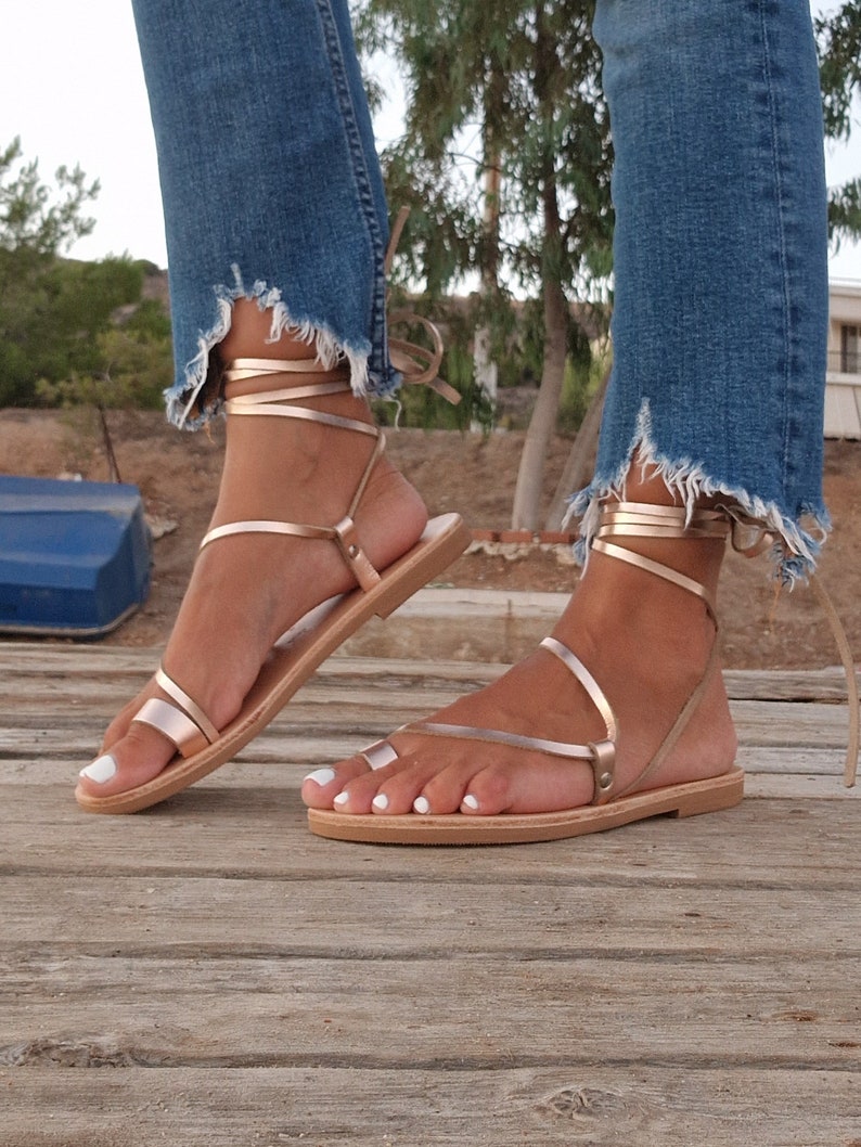May include: Rose gold sandals with thin straps and a flat sole. The sandals wrap around the ankle with a tie closure. The person is wearing blue jeans with a frayed hem. The sandals are on a wooden surface.