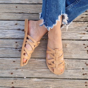 May include: A pair of tan leather sandals with a woven strap design. The sandals are worn on a wooden surface.
