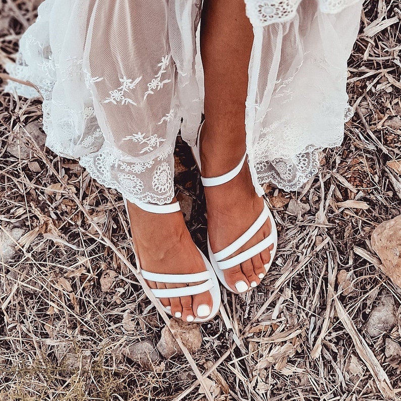 May include: A pair of white leather sandals with thin straps. The sandals are worn by a person wearing a white lace dress.