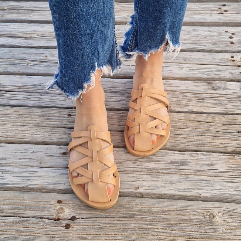 May include: Tan leather sandals with a woven design. The sandals are flat and have a simple, minimalist style.