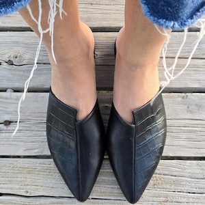 May include: A top-down view of two black leather mules with a pointed toe and a crocodile-textured vamp. The shoes are on a wooden deck, with the frayed edges of denim jeans visible at the top.