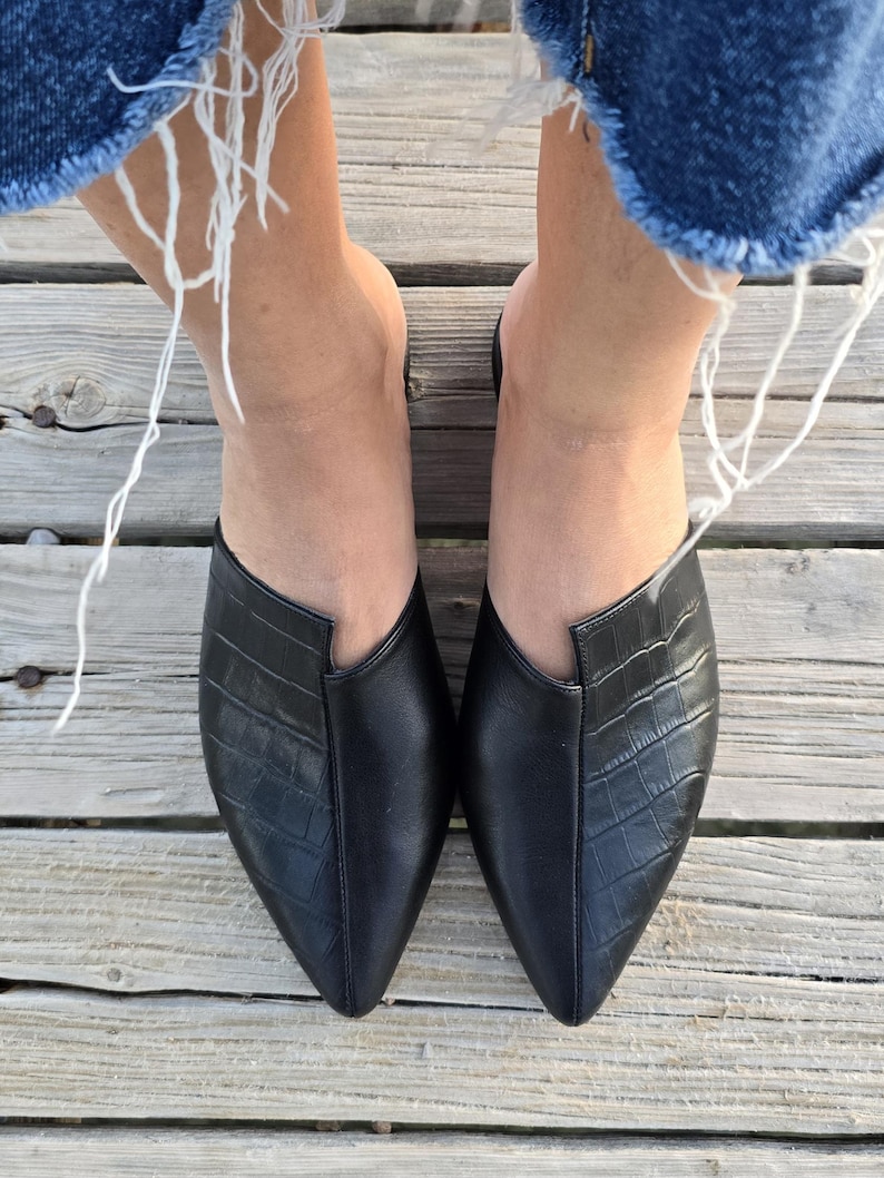 May include: A top-down view of two black leather mules with a pointed toe and a crocodile-textured vamp. The shoes are on a wooden deck, with the frayed edges of denim jeans visible at the top.