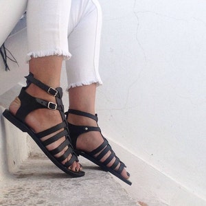 May include: Black leather gladiator sandals with multiple straps and buckles. The sandals have a flat sole and are worn on a person's feet.