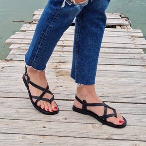 Op de afbeelding: Zwarte leren sandalen met gekruiste banden en een gespsluiting. De sandalen worden gedragen op een houten pier.