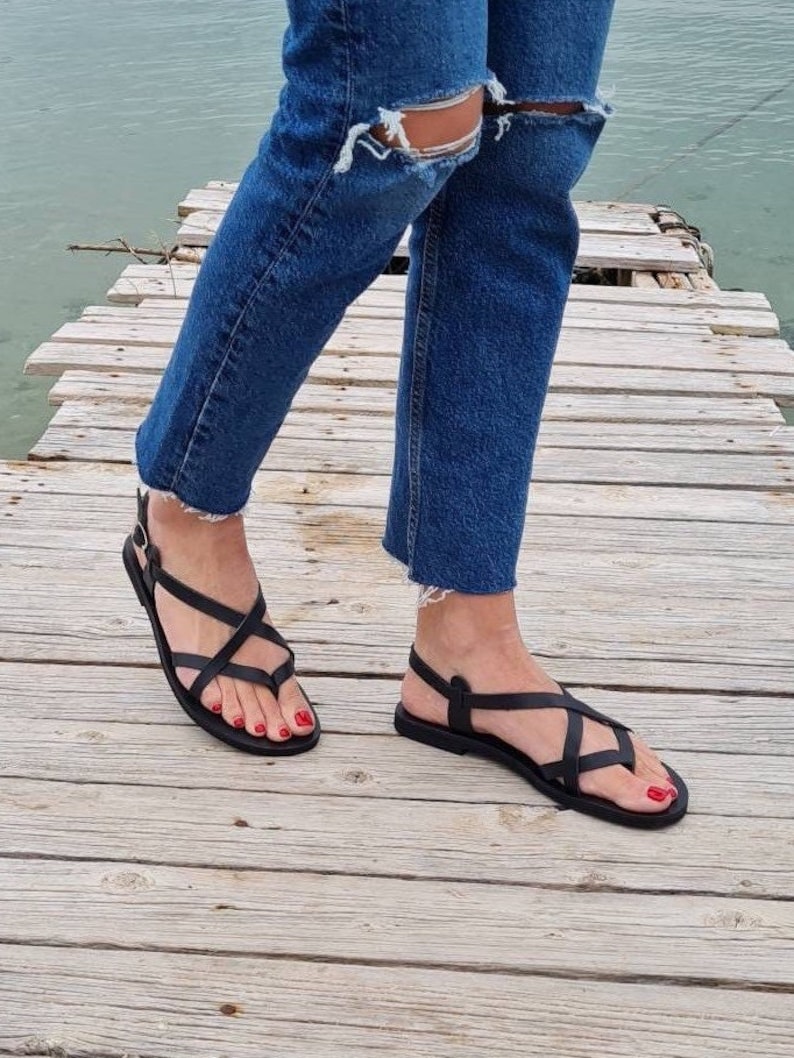 Op de afbeelding: Zwarte leren sandalen met gekruiste banden en een gespsluiting. De sandalen worden gedragen op een houten pier.