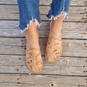May include: Tan leather sandals with a woven strap design. The sandals are worn on a wooden surface.