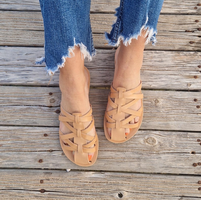 May include: Tan leather sandals with a woven strap design. The sandals are worn on a wooden surface.