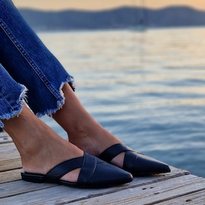 May include: A pair of black leather mules with a criss-cross strap design. The shoes have a pointed toe and a low heel. The shoes are worn by a person sitting on a wooden dock.