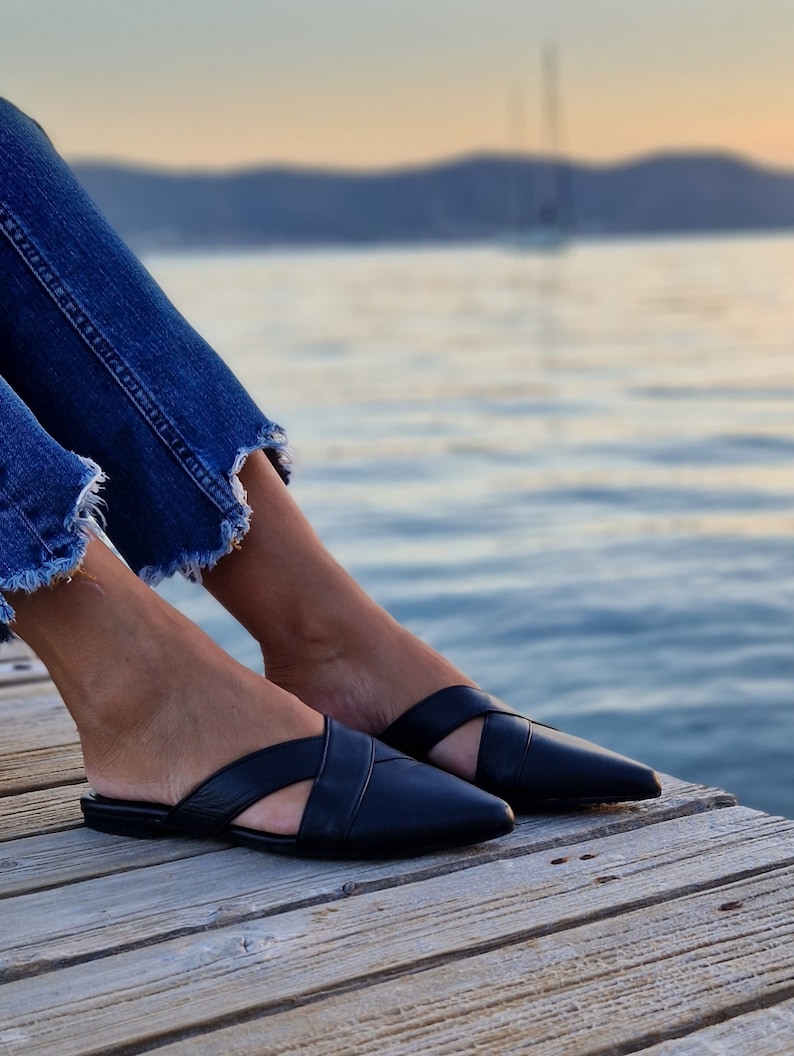 May include: A pair of black leather mules with a criss-cross strap design. The shoes have a pointed toe and a low heel. The shoes are worn by a person sitting on a wooden dock.