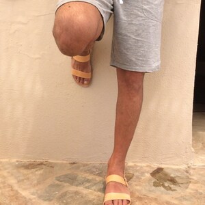 May include: A pair of tan leather sandals with two straps. The sandals are worn by a person standing on a stone floor.