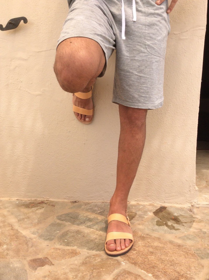 May include: A pair of tan leather sandals with two straps. The sandals are worn by a person standing on a stone floor.