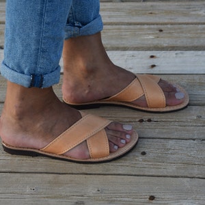 May include: A pair of tan leather sandals with a criss-cross strap design. The sandals are worn on a person's feet, with the toes visible.