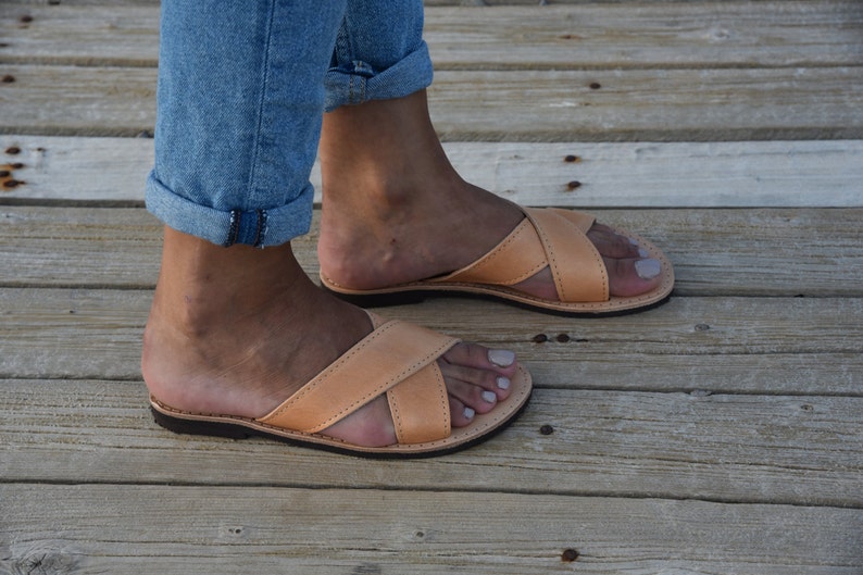 May include: A pair of tan leather sandals with a criss-cross strap design. The sandals are worn on a person's feet, with the toes visible.