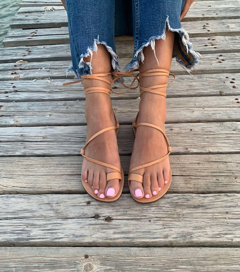 May include: Tan leather sandals with wrap-around ankle straps. The sandals have a toe loop and are worn with distressed denim jeans. The wooden deck provides a neutral background, highlighting the footwear.
