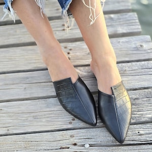 May include: Black leather slip-on shoes with a pointed toe and a crocodile texture. The shoes are worn with blue denim jeans with distressed hems. The shoes are on a wooden surface.