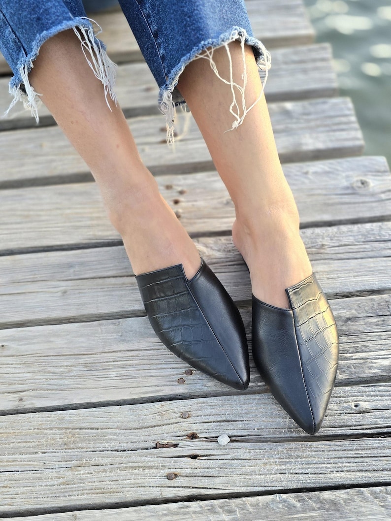 May include: Black leather slip-on shoes with a pointed toe and a crocodile texture. The shoes are worn with blue denim jeans with distressed hems. The shoes are on a wooden surface.