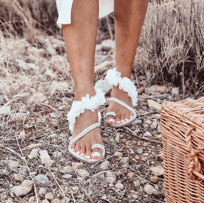 May include: White sandals with flower embellishments and pearl accents. The sandals have a toe ring and an ankle strap.
