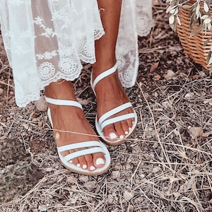 May include: White leather sandals with multiple straps. The sandals have a flat sole and are worn on a person's feet.