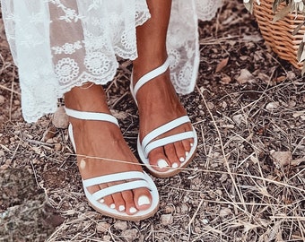 May include: White leather sandals with multiple straps. The sandals have a flat sole and are worn on a person's feet.