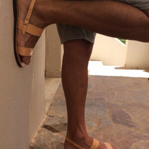 May include: Tan leather sandals with two straps and a buckle closure. The sandals are worn on a person's feet.