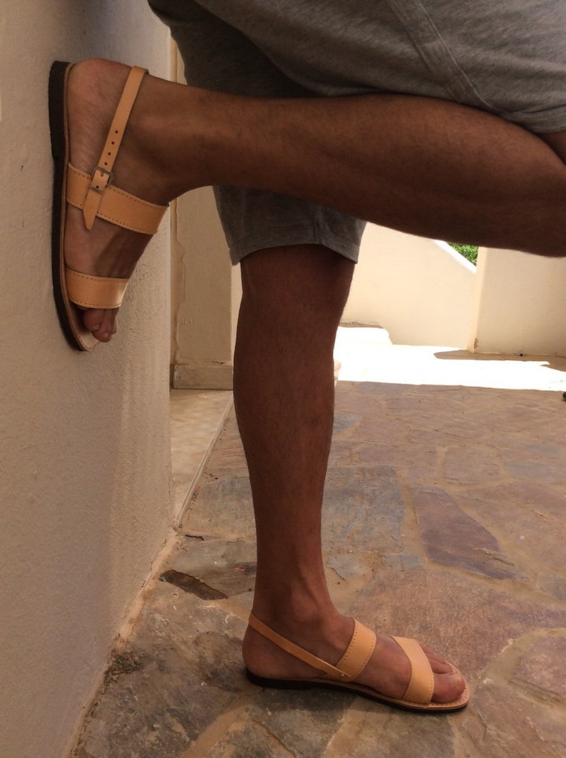May include: Tan leather sandals with two straps and a buckle closure. The sandals are worn on a person's feet.