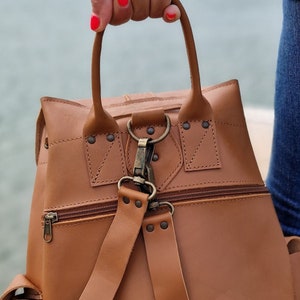 Puede incluir: Una mochila de cuero marr&oacute;n con asa superior y correas ajustables. La mochila tiene un bolsillo con cremallera en la parte delantera y un cierre de solapa con hebilla.