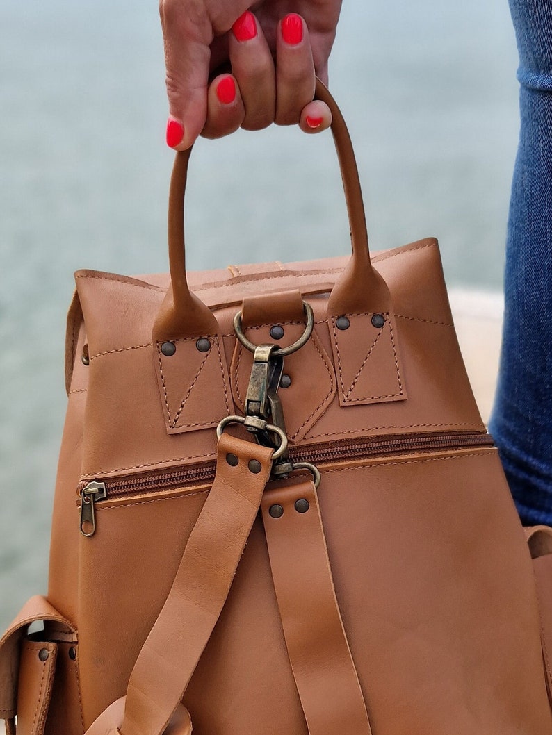 Puede incluir: Una mochila de cuero marr&oacute;n con asa superior y correas ajustables. La mochila tiene un bolsillo con cremallera en la parte delantera y un cierre de solapa con hebilla.
