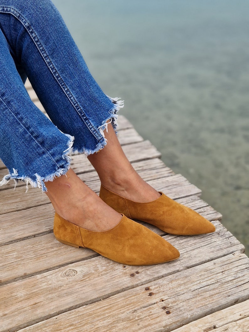 May include: A pair of brown suede pointed-toe flats on a wooden surface. The shoes are worn by a person wearing blue jeans.