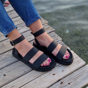 May include: Black leather sandals with two straps and an ankle strap with a buckle. The sandals have a platform sole.