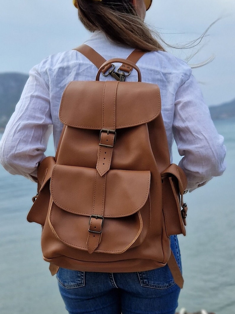 Puede incluir: Una mochila de cuero marr&oacute;n con dos bolsillos grandes en la parte delantera y un cierre de hebilla. La mochila tiene correas ajustables y un asa superior.