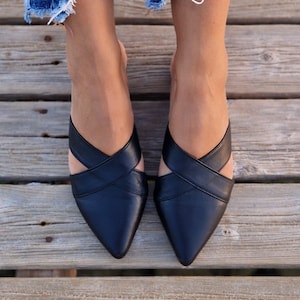 May include: A pair of black leather pointed-toe mules with criss-cross straps. The shoes are worn by a person with bare legs and denim shorts.