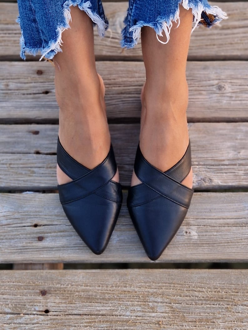 May include: A pair of black leather pointed-toe mules with criss-cross straps. The shoes are worn by a person with bare legs and denim shorts.