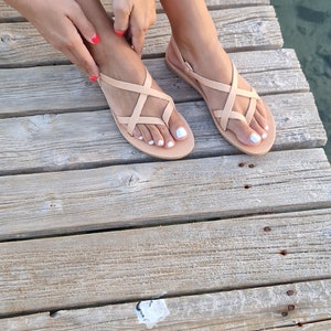 Op de afbeelding: Een paar lichtbruine leren sandalen met gekruiste banden. De sandalen worden gedragen door iemand die op een houten pier zit.