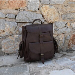 Puede incluir: Una mochila de cuero marr&oacute;n con varios bolsillos y cierre de hebilla. La mochila est&aacute; sobre una superficie de piedra.