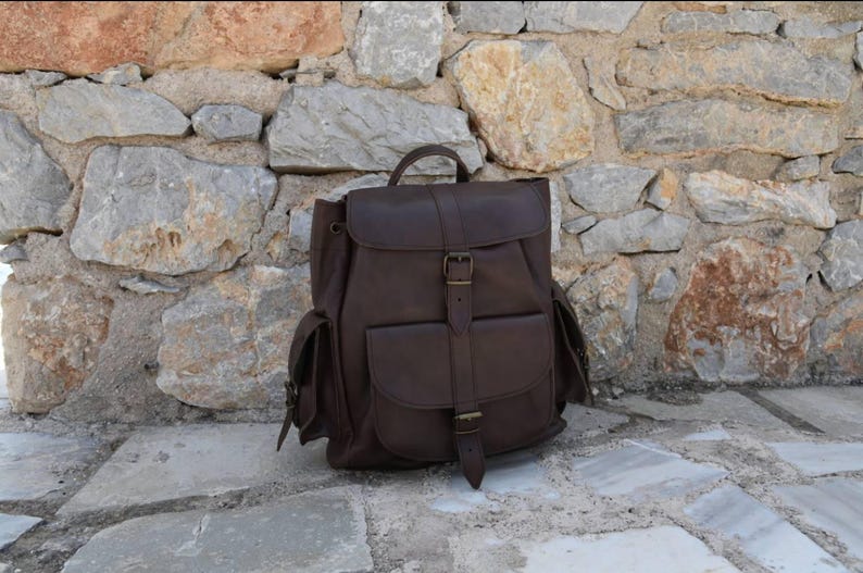 Puede incluir: Una mochila de cuero marr&oacute;n con varios bolsillos y cierre de hebilla. La mochila est&aacute; sobre una superficie de piedra.