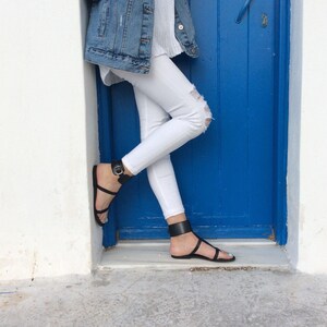 May include: A person wearing white jeans and a denim jacket with black ankle strap sandals. The person is standing in front of a blue door.