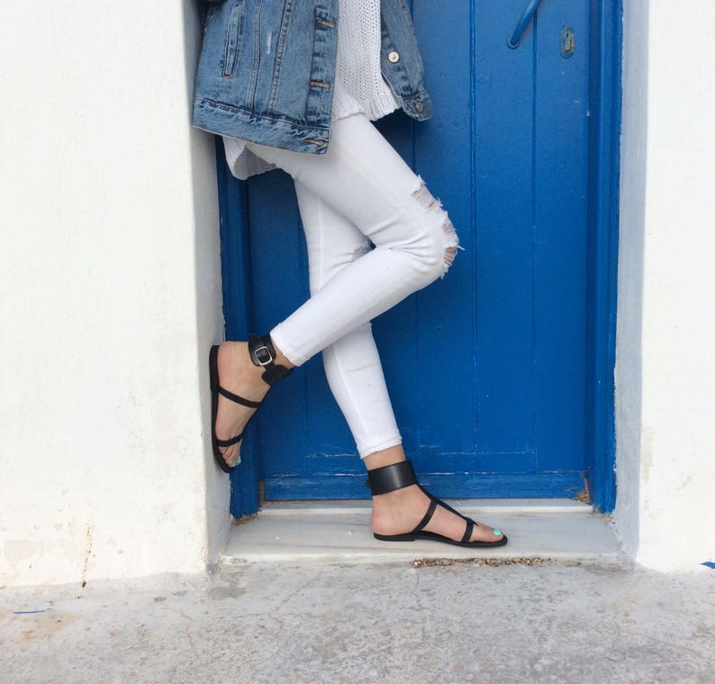 May include: A person wearing white jeans and a denim jacket with black ankle strap sandals. The person is standing in front of a blue door.