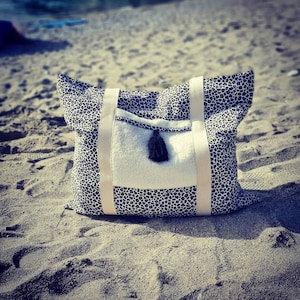 May include: A black and white leopard print tote bag with a white pocket and a black tassel. The bag is sitting on a sandy beach.