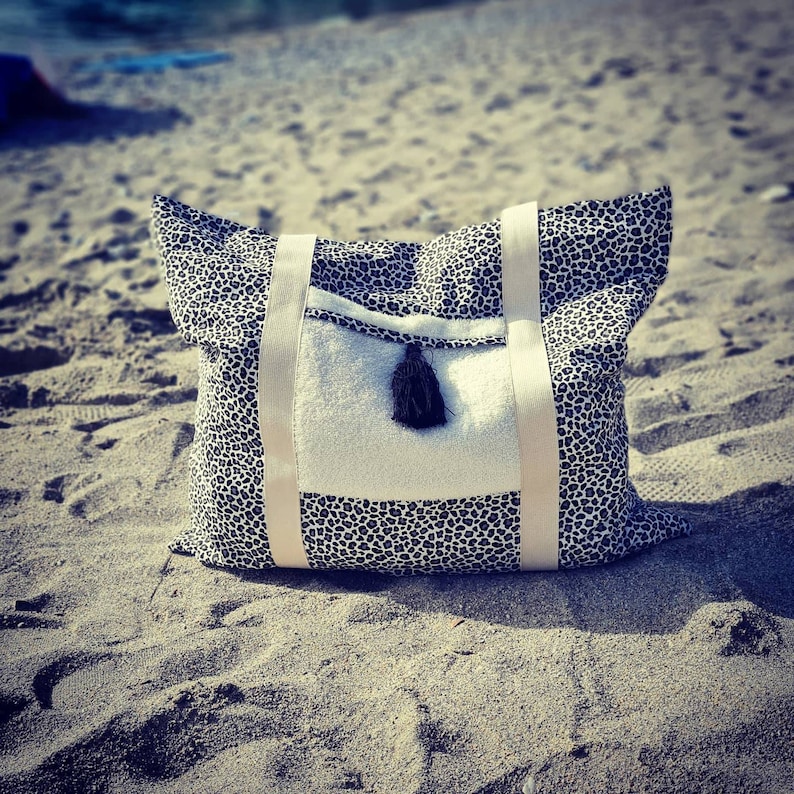 May include: A black and white leopard print tote bag with a white pocket and a black tassel. The bag is sitting on a sandy beach.
