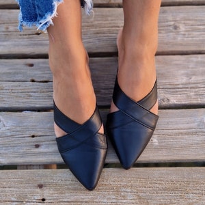 May include: A pair of black leather pointed-toe mules with criss-cross straps. The shoes are worn by a person wearing denim jeans.