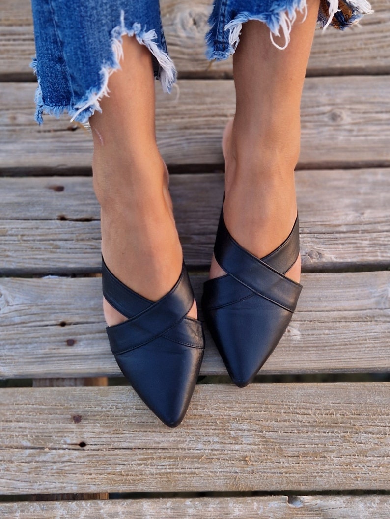 May include: A pair of black leather pointed-toe mules with criss-cross straps. The shoes are worn by a person wearing denim jeans.