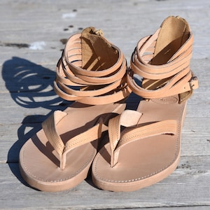 Può includere: Un paio di sandali in pelle color sabbia con più cinturini intorno alla caviglia. I sandali hanno un design semplice e una suola piatta.